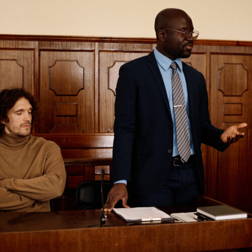 Confident African American male attorney in formalwear explaining points of case to judge while standing next to suspect during trial session